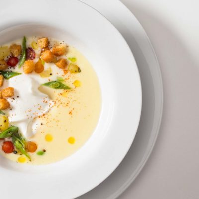 Cold Potato and Leek Soup Cold potato and leak soup with yogurt, green beans, potatoes, currant tomatoes, croutons, extra virgin olive oil and hot pepper. Served on a white ceramic bowl on a white background.